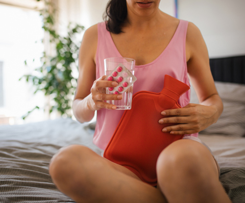 woman at home suffering from menstrual pain, taking painkillers, warming lower abdomen with a hot water bottle